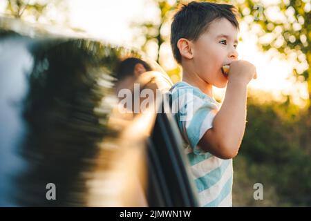 Petit garçon caucasien qui colle de la voiture et mange son snack tout en regardant la beauté de la nature. Bonne famille, enfance. Vacances d'été. Banque D'Images