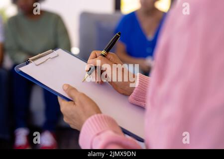 Section médiane de la femme thérapeute caucasienne âgée écrivant sur papier avec le presse-papiers dans la maison de soins infirmiers Banque D'Images