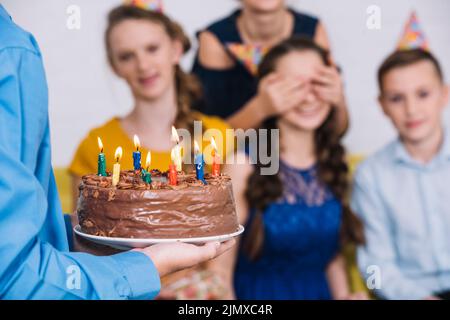 Gros plan la main de garçon apportant gâteau au chocolat anniversaire fille avec les yeux couverts par son ami Banque D'Images