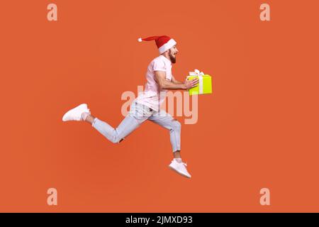 Homme barbu sautant dans l'air ou courant rapidement pour donner la boîte cadeau de Noël, exprimant le bonheur, portant un T-shirt rose et chapeau de père noël. Studio d'intérieur isolé sur fond orange. Banque D'Images