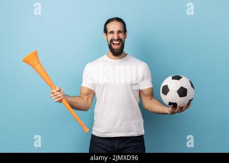 Portrait d'un homme joyeux avec une barbe portant un T-shirt blanc tenant une corne de taureau et un ballon de football, célébrant la victoire de l'équipe de football préférée. Studio d'intérieur isolé sur fond bleu. Banque D'Images