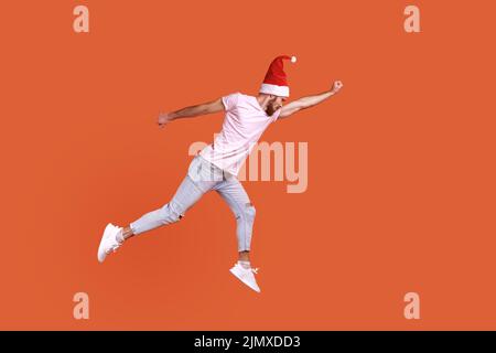 Pleine longueur d'homme à santa chapeau volant dans l'air en tant que super-héros avec la main levée, sensation de super puissance, étant pressé de célébrer le nouvel an, en portant un T-shirt. Studio d'intérieur isolé sur fond orange Banque D'Images