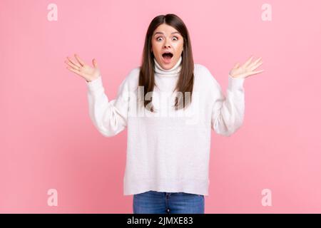 Portrait d'une jolie femme choquée avec des bras relevés exprimant l'étonnement, en gardant la bouche largement ouverte, en portant blanc pull de style décontracté. Studio d'intérieur isolé sur fond rose. Banque D'Images