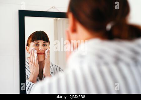 Avez-vous commencé votre voyage vers une belle peau. Une jeune femme attrayante appliquant un soin de beauté sur son visage en se tenant debout devant le Banque D'Images