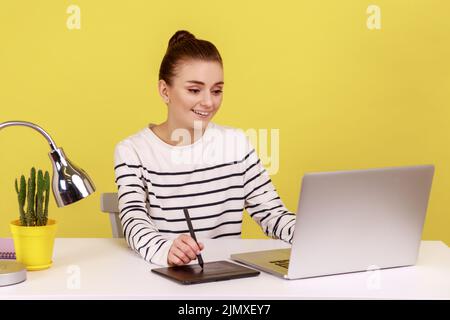 Belle femme souriante créatrice designer assis sur le lieu de travail avec tablette graphique et ordinateur portable, travaillant sur des équipements d'art numérique professionnels. Studio d'intérieur tourné isolé sur fond jaune. Banque D'Images