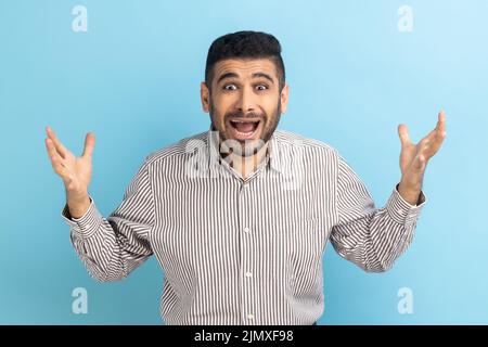 Un homme d'affaires agité avec la barbe a des problèmes, gesticuler avec les mains, crise et négativité, malentendu, porter une chemise rayée. Studio d'intérieur isolé sur fond bleu. Banque D'Images