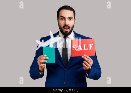 Homme barbu surpris tenant un passeport avec avion en papier et signe de vente, exprimant l'étonnement, voyager, portant un costume de style officiel. Prise de vue en studio isolée sur fond gris. Banque D'Images