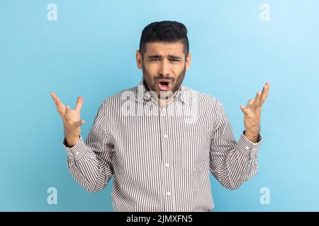 Agacé homme d'affaires stressé avec la barbe de demander ce que vous voulez et de se répandre les mains, querelle de famille, malentendu, porter une chemise rayée. Studio d'intérieur isolé sur fond bleu. Banque D'Images