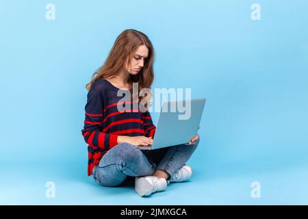 Portrait d'une femme bouleversée portant un chandail rayé de style décontracté, assis tenant le carnet à genoux, regardant l'écran, sensation de tristesse. Studio d'intérieur isolé sur fond bleu. Banque D'Images