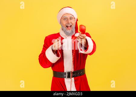 Homme âgé avec barbe grise portant le costume du père noël tenant le combiné téléphonique fixe, vous appelant à féliciter avec des commodes, pointant vers l'appareil photo. Studio d'intérieur isolé sur fond jaune Banque D'Images