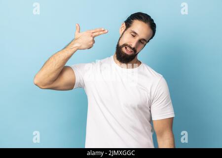 Coup de tête, tuez-moi s'il vous plaît. Homme déprimé avec la barbe portant un T-shirt blanc se suicider avec un geste de doigt de pistolet, expression de visage désespérée. Studio d'intérieur isolé sur fond bleu. Banque D'Images