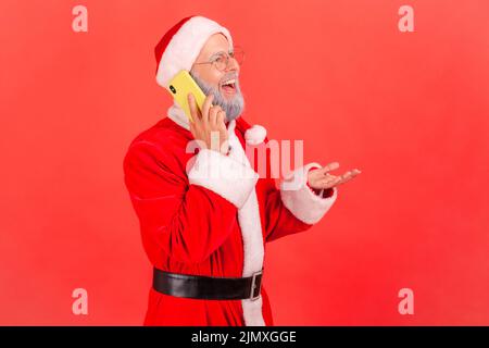 Joyeux homme âgé souriant avec barbe grise portant le costume du père noël téléphone parlant, parlant avec expression positive, nouvelles agréables. Studio d'intérieur isolé sur fond rouge. Banque D'Images