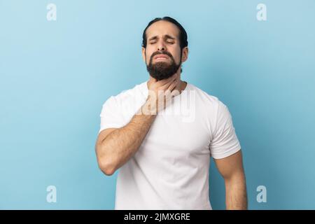 Portrait d'un homme malsain avec une barbe portant un T-shirt blanc souffrant de maux de gorge, gardant la main sur son cou, froissant le visage et les yeux fermés. Studio d'intérieur isolé sur fond bleu. Banque D'Images