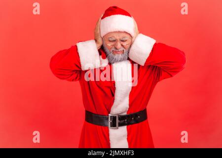 Je ne veux pas ici. Homme âgé avec barbe grise portant le costume du père noël debout couvrant les oreilles avec les paumes, bruit fort, visage froid. Studio d'intérieur isolé sur fond rouge. Banque D'Images