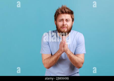 S'il vous plaît, je suis mendiant. Portrait de l'homme barbu tenant les bras dans la prière, demandant l'aide ou le pardon avec les yeux implorants, sincère demandant la permission. Studio d'intérieur isolé sur fond bleu. Banque D'Images
