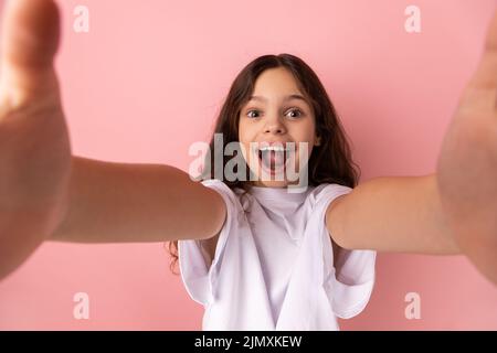 Portrait d'une petite fille choquée portant un T-shirt blanc prenant selfie, regardant l'appareil photo avec la bouche ouverte POV, point de vue de la photo. Studio d'intérieur isolé sur fond rose. Banque D'Images