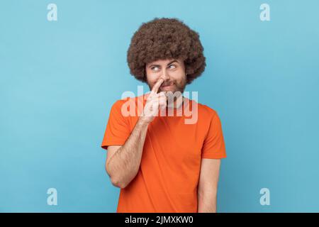 Portrait de l'homme puéril avec une coiffure afro portant un T-shirt orange cueillant le nez et collant hors de la langue avec l'expression stupide comique. Studio d'intérieur isolé sur fond bleu. Banque D'Images