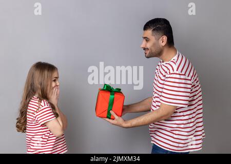 Vue latérale du père donnant une boîte cadeau rouge à sa fille, la famille en T-shirts à rayures, la petite fille excitée étant heureuse d'obtenir un cadeau à son anniversaire. Prise de vue en studio isolée sur fond gris. Banque D'Images