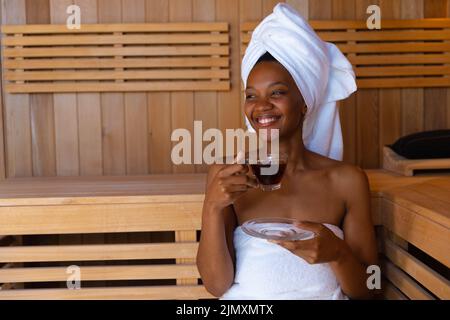 Jeune femme afro-américaine joyeuse regardant loin tout en ayant une boisson saine dans le sauna, l'espace de copie Banque D'Images