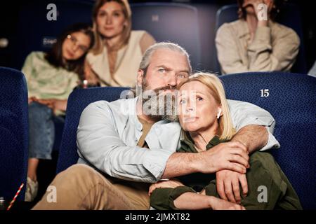 Homme caucasien adulte mature avec une barbe sur le visage qui embrasse sa femme tout en regardant un triste film dramatique au cinéma Banque D'Images