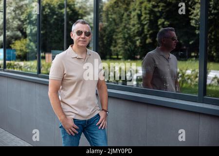 Portrait d'un homme brutal senior en lunettes de soleil marchant avec les mains dans les poches de jeans le long de l'immeuble de bureau moderne, reflet de silhouette dans la fenêtre Banque D'Images