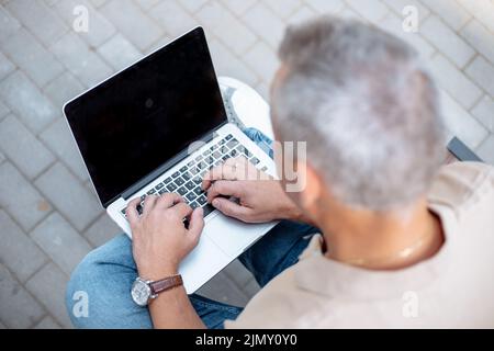 Vue sur le dessus photo d'un homme d'affaires aux cheveux gris, assis sur un banc dans la rue de la ville et dactylographiant sur un ordinateur portable numérique, écran noir. Style de vie moderne, travail à distance Banque D'Images