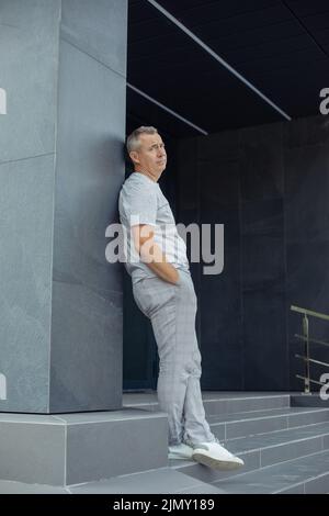 Portrait d'un homme d'affaires vieux et fatigué aux cheveux gris penché en arrière sur la construction grise d'un immeuble de bureaux moderne près de l'entrée. Style de vie urbain, élégant Banque D'Images