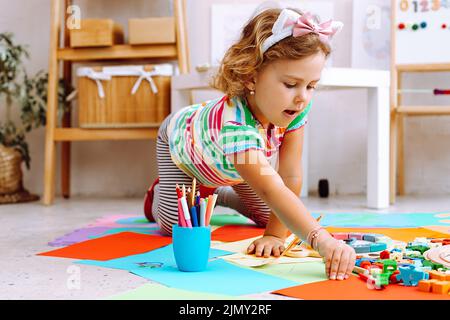 Petite fille avec beau cadre sur la tête assis sur le plancher et atteignant pour la feuille de papier dans la maternelle. Dessin avec des crayons de couleur, leçon intéressante Banque D'Images