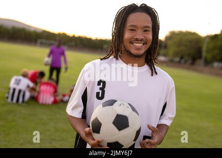 Portrait d'un homme biracial souriant en uniforme blanc tenant le ballon de football debout sur l'aire de jeux Banque D'Images