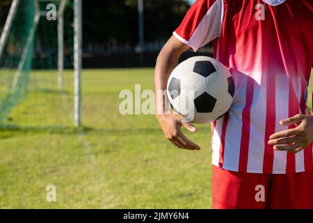 Section médiane d'un joueur masculin caucasien portant un maillot rouge avec un ballon de football debout sur le terrain de jeu Banque D'Images