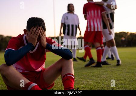 Joueur de football multiracial triste masculin avec la tête dans les mains assis sur terre herbeuse contre ciel clair Banque D'Images