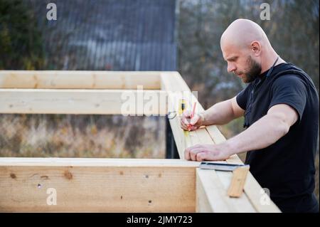 Homme ouvrier bâtiment maison de cadre en bois sur la base de pile. Charpentier à tête blanche à l'aide d'un mètre à ruban pour mesurer des planches en bois et faire des marques avec un crayon. Concept de menuiserie. Banque D'Images