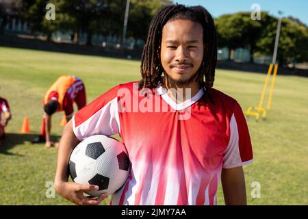 Portrait d'un homme souriant biracial avec des dreadlocks tenant le ballon de football sur l'aire de jeux en été Banque D'Images