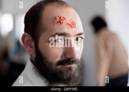 Portrait d'un homme avec une plaie zombie sanglante, artiste préparant un maquillage et des effets spéciaux pour transformer une personne en cadavre infecté. Concevoir un costume d'horreur créepy avec le visage décédé. Banque D'Images