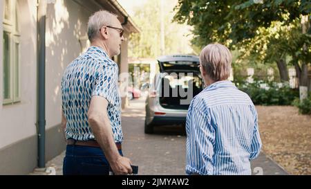 Couple senior mettant des bagages dans le coffre de voiture tout en se prépare pour les vacances. Des aînés joyeux se rendent à la retraite tout en ayant de lourds bagages et un chariot de voyage. Banque D'Images