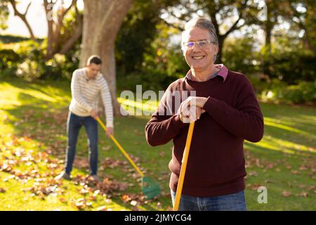 Image d'un père caucasien heureux et d'un fils adulte qui balaie les feuilles dans le jardin Banque D'Images