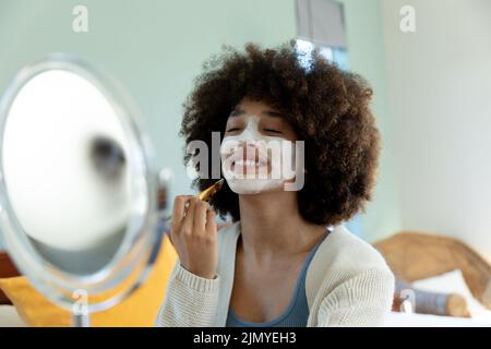 Jeune femme biraciale souriante avec cheveux afro appliquant de la crème de beauté sur le visage tout en regardant le miroir Banque D'Images