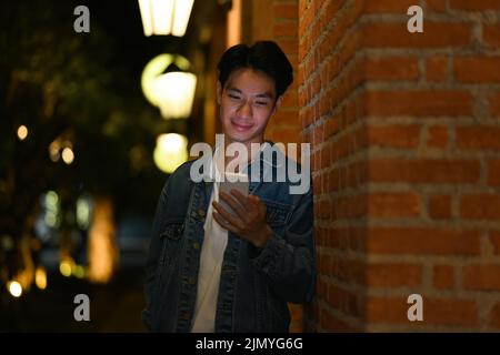 Portrait d'un homme asiatique élégant qui tape du texte sur un smartphone tout en étant debout dans les rues nocturnes de la ville Banque D'Images