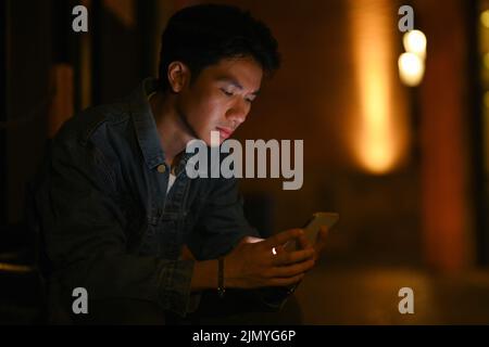 Jeune homme hippster utilisant un téléphone mobile, bavardant avec des amis sur les réseaux sociaux tout en étant assis dans les rues de la ville le soir Banque D'Images