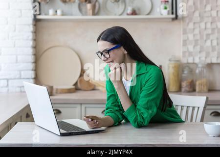 Achats en ligne. Une jeune belle femme en lunettes et une chemise verte est assise à la maison avec un ordinateur portable, tient une carte de crédit dans ses mains. Effectue des achats sur Internet, commande la livraison, paie en ligne. Banque D'Images