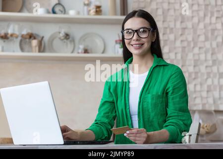 Achats en ligne. Portrait d'une belle femme en lunettes et une chemise verte est assise à la maison avec un ordinateur portable, tient une carte de crédit dans ses mains. Effectue des achats sur Internet, commande la livraison, paie en ligne. Banque D'Images