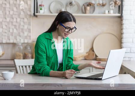 Échec des achats en ligne. La banque a bloqué le compte sur la carte de crédit détenue par une jeune femme en lunettes et en chemise verte. Elle est assise avec un ordinateur portable à la maison, contrariée, confuse, elle se répand les mains. Banque D'Images