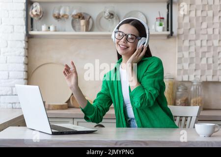 Portrait d'une jeune femme écoutant de la musique dans des écouteurs blancs, des lunettes et des vêtements verts. Elle s'assoit à la table à la maison avec un ordinateur portable, ferme les yeux, danse, aime Banque D'Images