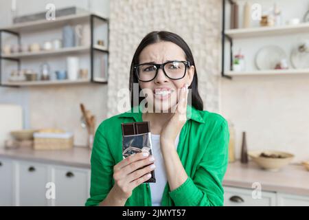 Mal de dents. Portrait d'une jeune belle femme à la maison en lunettes et une chemise verte qui a un mal de dents de bonbons. Il tient une barre de chocolat dans ses mains, tient sa joue, ressent une douleur sévère. Banque D'Images