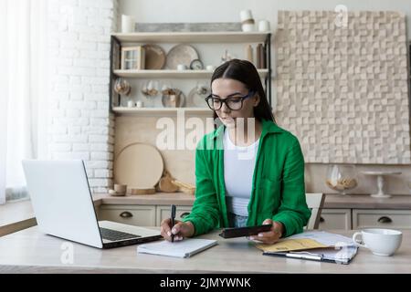 La comptabilité à domicile.Une jeune femme compte son revenu et expenses.checks le budget du ménage. En lunettes et une chemise verte, se trouve à la maison avec un ordinateur portable, une calculatrice et écrit dans un bloc-notes, calcule. Banque D'Images