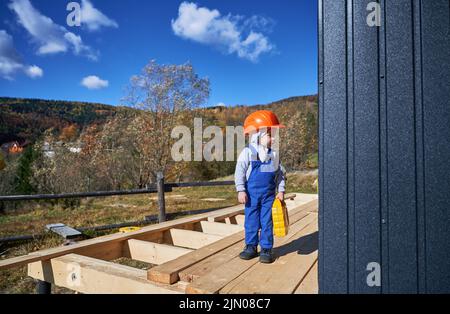 Garçon tout-petit jouant comme constructeur sur le chantier de construction. Menuisier d'enfant en casque orange et combinaison bleue apprenant à construire la maison de cadre en bois à l'extérieur le jour ensoleillé. Conception de la menuiserie et de l'atelier. Banque D'Images