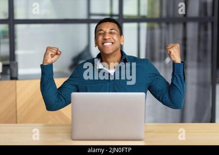Un jeune homme enthousiaste qui célèbre son succès tout en étant assis sur un lieu de travail équipé d'un ordinateur portable, un homme d'affaires heureux qui a de bonnes nouvelles Banque D'Images