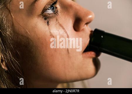 La jeune femme qui pleure avec du mascara étalé boit de l'alcool du cou de la bouteille. Portrait de la vue latérale d'une femme malheureuse qui connaît une crise émotionnelle dans la vie Banque D'Images