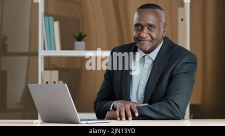 Portrait d'un homme d'affaires africain mature et âgé assis au bureau à table avec un ordinateur portable qui hoche la tête en répondant oui à l'approbation en faveur de la bonne idée Banque D'Images