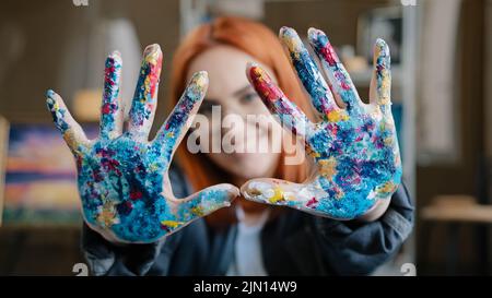 Gros plan heureuse femme funy avec les cheveux rouges peintre fille artiste en lunettes regarde la caméra étire les mains vers l'avant montrant les paumes sales tachées dans l'acrylique Banque D'Images